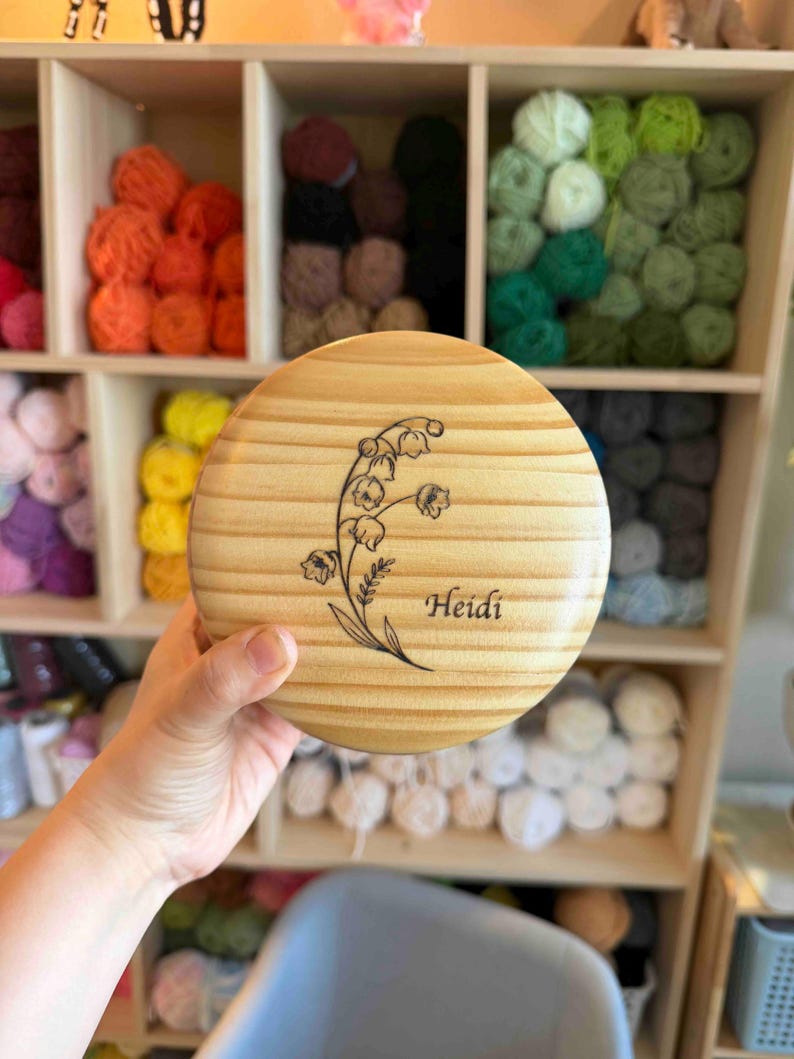May include: A round, wooden box lid with a floral design and the name "Heidi" engraved on it. The lid is light brown with visible wood grain. The background shows shelves filled with colorful yarn balls.