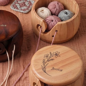 May include: Wooden yarn bowls with yarn inside. One bowl is open, displaying several balls of yarn in shades of pink, gray, and cream. The other bowls are closed, with the name "Jessica" engraved on the lid. A crocheted square is in the background.