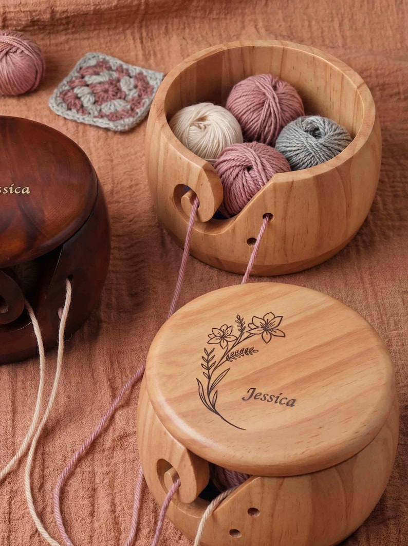 May include: Wooden yarn bowls with yarn inside. One bowl is open, displaying several balls of yarn in shades of pink, gray, and cream. The other bowls are closed, with the name "Jessica" engraved on the lid. A crocheted square is in the background.