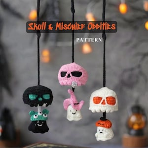 May include: Three hanging crochet ornaments featuring skulls and small figures. The left is a black skull with a green creature, the center is a pink skull with a white ghost wearing a pink hat, and the right is a white skull with an orange-topped ghost.