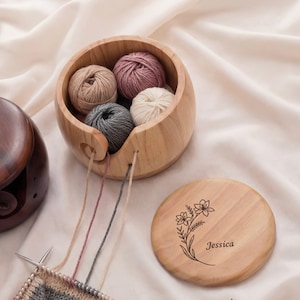 May include: A wooden yarn bowl with several yarn balls in shades of pink, beige, gray, and white. The bowl has a slot for the yarn to feed through. A wooden lid with a floral design and the name "Jessica" is next to the bowl. Knitting needles and yarn are in the foreground.