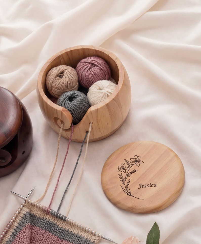May include: A wooden yarn bowl with several yarn balls in shades of pink, beige, gray, and white. The bowl has a slot for the yarn to feed through. A wooden lid with a floral design and the name "Jessica" is next to the bowl. Knitting needles and yarn are in the foreground.