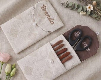 May include: Two cream-colored fabric cases with a diamond-quilted pattern and a single white button closure. One case is embroidered with the name Brenda. The other case holds five brown crochet hooks and black scissors.