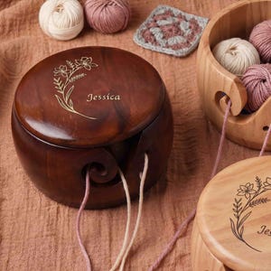 May include: A wooden yarn bowl with a floral design and the name "Jessica" engraved on the lid. The bowl is dark brown and has a swirl-shaped yarn guide. Several balls of yarn in cream and pink hues are visible, along with a small crocheted square.