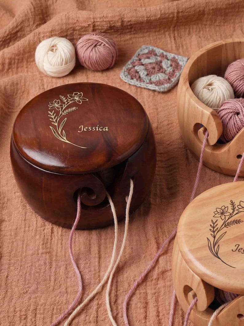 May include: A wooden yarn bowl with a floral design and the name "Jessica" engraved on the lid. The bowl is dark brown and has a swirl-shaped yarn guide. Several balls of yarn in cream and pink hues are visible, along with a small crocheted square.