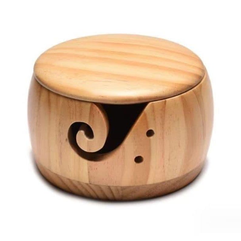 May include: A light-colored wooden yarn bowl with a lid. The bowl has a rounded shape with a swirl-shaped opening on the side for the yarn to pass through. Two small holes are also visible. The lid sits on top of the bowl.