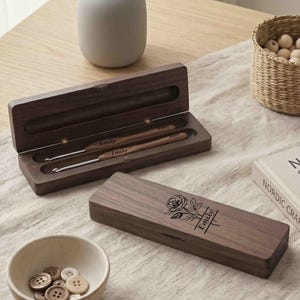 Puede incluir: Juego de ganchillos de madera en un estuche marrón oscuro. El estuche está grabado con una rosa y el nombre "Emily". El juego incluye dos ganchillos. También se ve un cuenco pequeño con botones y un jarrón decorativo.