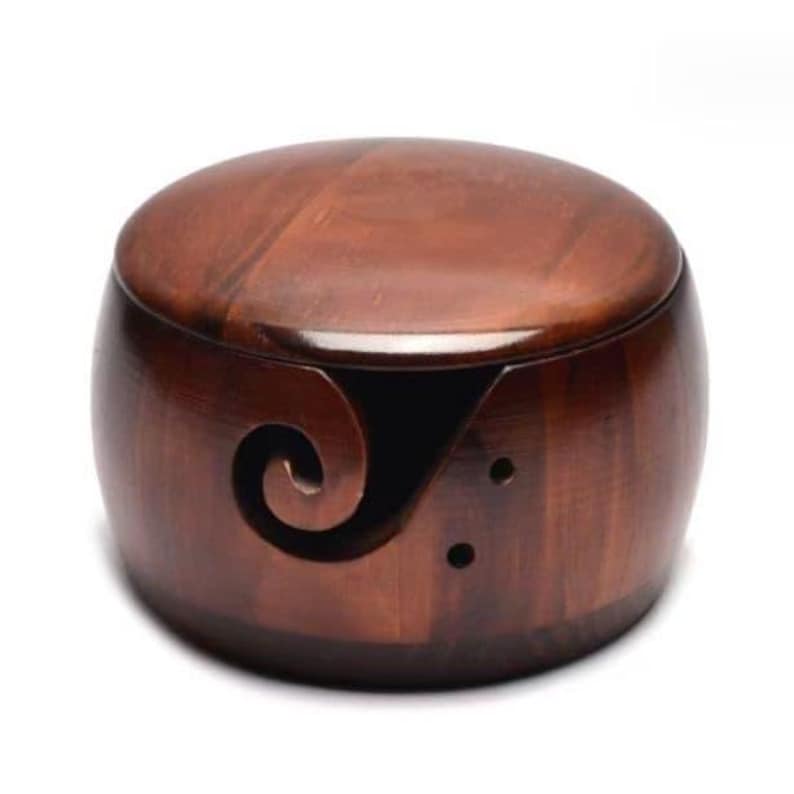 May include: A wooden yarn bowl with a lid. The bowl is a rich brown color and has a smooth, polished finish. A decorative swirl is carved into the side of the bowl, and there are two small holes. The bowl is designed to hold yarn while knitting or crocheting.