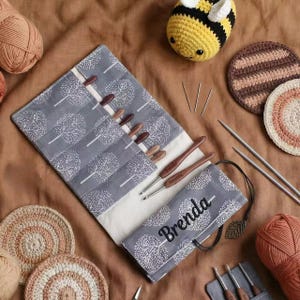 May include: A gray fabric roll-up case with a white tree pattern, personalized with the name Brenda in black script. It contains wooden crochet hooks, with two hooks engraved with the names Emily and Amanda, alongside yarn balls and coasters.
