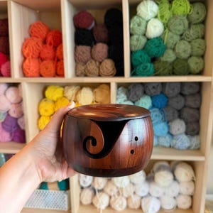 May include: A wooden yarn bowl with a dark brown finish, held in a hand. The bowl has a curved slot for yarn and two small holes. Shelves in the background are filled with colorful yarn balls, showcasing a craft supply theme.