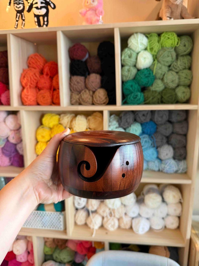 May include: A wooden yarn bowl with a dark brown finish, held in a hand. The bowl has a curved slot for yarn and two small holes. Shelves in the background are filled with colorful yarn balls, showcasing a craft supply theme.