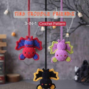 May include: Three hanging crochet bat ornaments labeled TINY TROUBLE FRIENDS 3-IN-1 Crochet Pattern. One bat is red and blue, one is purple and light green, and one is black and yellow. Each bat has small black eyes and hangs from a colored cord.