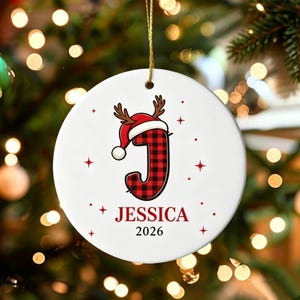 May include: A round white ceramic Christmas ornament featuring a red and black buffalo plaid letter J wearing a Santa hat with antlers. The name JESSICA and the year 2026 are printed in red and black text below the letter, surrounded by red stars.