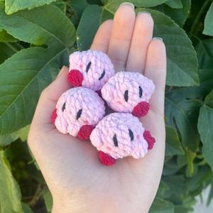 Puede incluir: Cuatro figuras de ganchillo hechas a mano con forma de personaje rosa con pies rojos y cara negra. Las figuras se sostienen en una mano sobre un fondo de hojas verdes. Las figuras son pequeñas y detalladas.