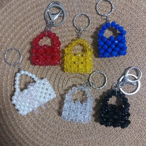 Könnte beinhalten: Eine Sammlung von Schlüsselanhängern mit Perlenhandtaschen in verschiedenen Farben, darunter Rot, Gelb, Blau, Weiß, Klar und Schwarz. Jeder Schlüsselanhänger hat ein kleines, handtaschenförmiges Design mit einem silberfarbenen Schlüsselring.