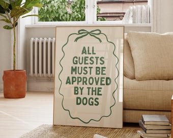 May include: A framed poster with the text "ALL GUESTS MUST BE APPROVED BY THE DOGS" in green lettering. The poster is on a beige rug in a living room setting, with a sofa, wooden chair, and a potted plant.