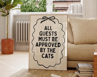 May include: A framed print with the text "ALL GUESTS MUST BE APPROVED BY THE CATS" in a living room setting. The print is on a rug in front of a window. A wooden chair and a potted plant are also in the room. The room has light-colored walls and flooring.