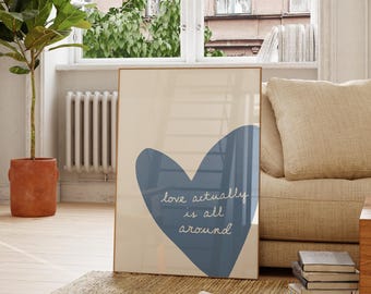 May include: A framed print with a blue heart and the words "love actually is all around" in white script. The print is leaning against a beige sofa in a well-lit room with a wooden chair and a potted plant.