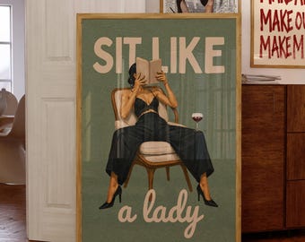 May include: A framed wall art print featuring a sage green background with the text "SIT LIKE a lady" in cream. The illustration depicts a person in a black lace top and wide-leg trousers sitting in a chair, reading a book with a glass of red wine.