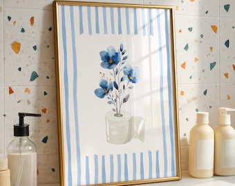 May include: A framed watercolor print featuring blue flowers and a roll of toilet paper. The artwork is set against a blue and white striped background, encased in a gold frame. The image is displayed on a white surface.