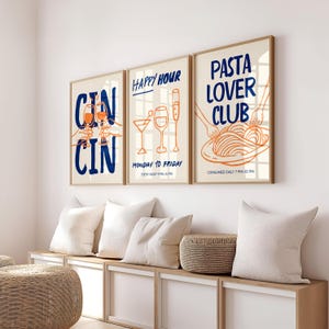 May include: Three framed art prints with a beige background and wooden frames. The prints feature illustrations and text in blue and orange. The first says "CIN CIN", the second "HAPPY HOUR", and the third "PASTA LOVER CLUB".