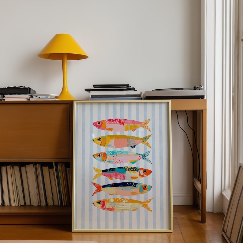 May include: A framed vertical art print featuring six stylized fish stacked in a column against a light blue and white vertical striped background. Each fish is rendered in a different color palette including pink, yellow, teal, and orange with patterns.