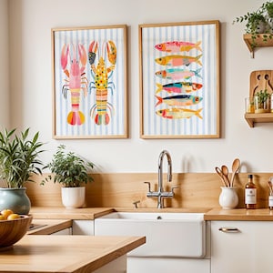 May include: Two framed wall art prints featuring colorful marine life illustrations. The left print displays two stylized lobsters, while the right print shows a vertical stack of six multi-colored fish, both set against a light blue striped background.