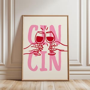 Puede incluir: Una impresión artística enmarcada con un fondo crema y texto rosa que dice "CIN CIN". Dos copas de vino tinto chocan, sostenidas por manos delineadas en rojo. La obra de arte es de estilo minimalista.