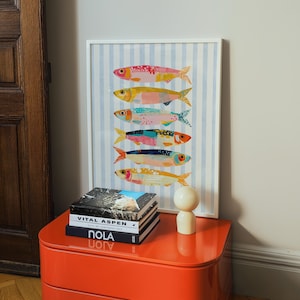 May include: A framed wall art print featuring six stylized fish stacked vertically, each with unique colorful patterns, set against a light blue and white vertical striped background. The print is housed in a thin white frame.