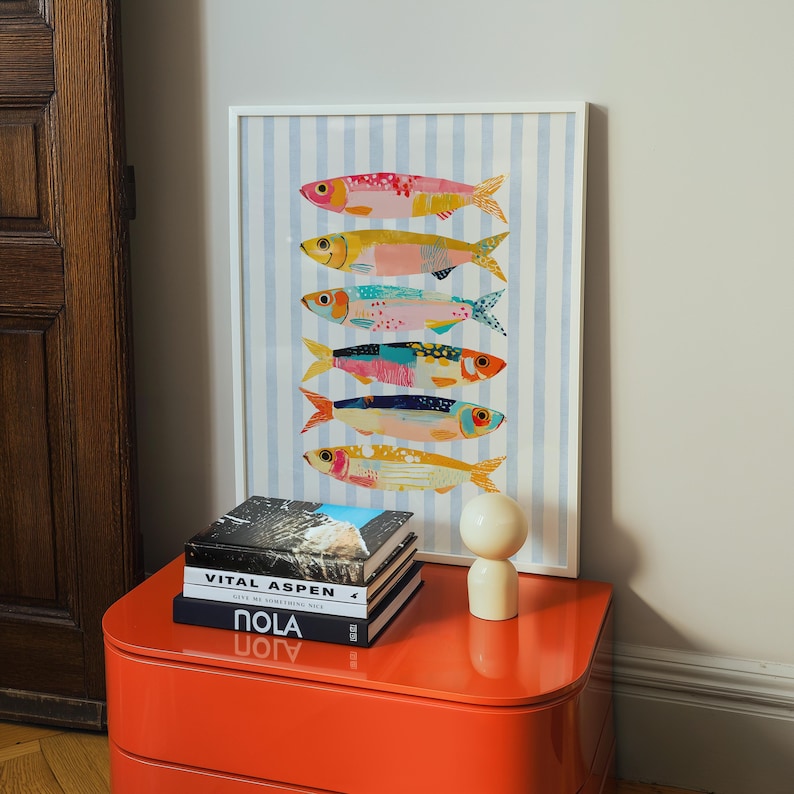 May include: A framed wall art print featuring six stylized fish stacked vertically, each with unique colorful patterns, set against a light blue and white vertical striped background. The print is housed in a thin white frame.