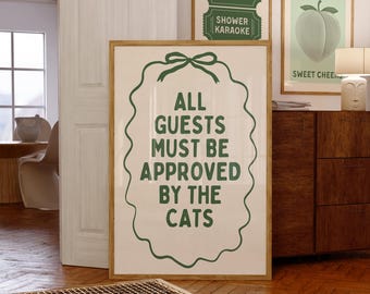 May include: A framed print with the text "ALL GUESTS MUST BE APPROVED BY THE CATS" in green. Two smaller framed prints read "SHOWER KARAOKE" and "TAKE A SEAT". The artwork is displayed in a room with wooden floors.