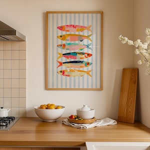 May include: A framed vertical wall art print featuring six stylized fish stacked in a column against a light blue and white vertical striped background. The fish are rendered in various bright colors including pink, yellow, teal, and orange.