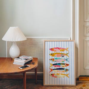 May include: A framed vertical print featuring six stylized fish stacked horizontally, each with unique colorful patterns, set against a light blue and white vertical striped background. The artwork is displayed in a light wood frame.