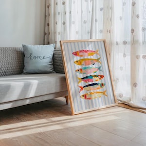 May include: A framed vertical art print featuring six stylized fish stacked in a column against a light blue and white vertical striped background. Each fish is rendered in a different color palette, including shades of pink, yellow, teal, orange, and blue, with distinct patterns and textures on their bodies. The frame is a light-toned wood.
