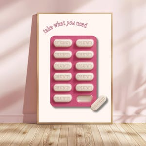 Op de afbeelding: Een ingelijste print met een roze blisterverpakking met pillen, elk met het woord "CREATIVITY". Een losse pil, ook met "CREATIVITY", ligt ernaast. De tekst "take what you need" staat erboven. Gouden lijst, roze muur en houten vloer.
