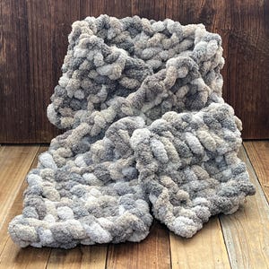 May include: A chunky knit scarf in shades of gray, white, and tan. The scarf is made of a thick, soft yarn, and is folded over on a wooden surface. The texture of the yarn is visible, creating a cozy and warm appearance.