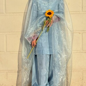 May include: A light blue salwar kameez outfit with a sheer dupatta. The outfit includes a tunic top and loose-fitting pants. The dupatta has gold trim. A person is holding a sunflower.