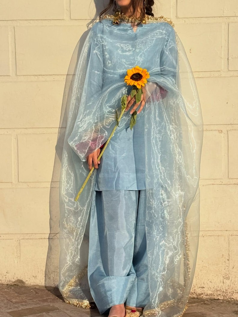 May include: A light blue salwar kameez outfit with a sheer dupatta. The outfit includes a tunic top and loose-fitting pants. The dupatta has gold trim. A person is holding a sunflower.