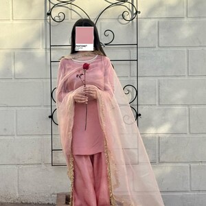 May include: A light pink salwar kameez outfit. The ensemble includes a long-sleeved tunic, wide-legged pants, and a sheer dupatta with gold trim. A red rose is held in the hands. The background features a decorative black metal arch and a white brick wall.
