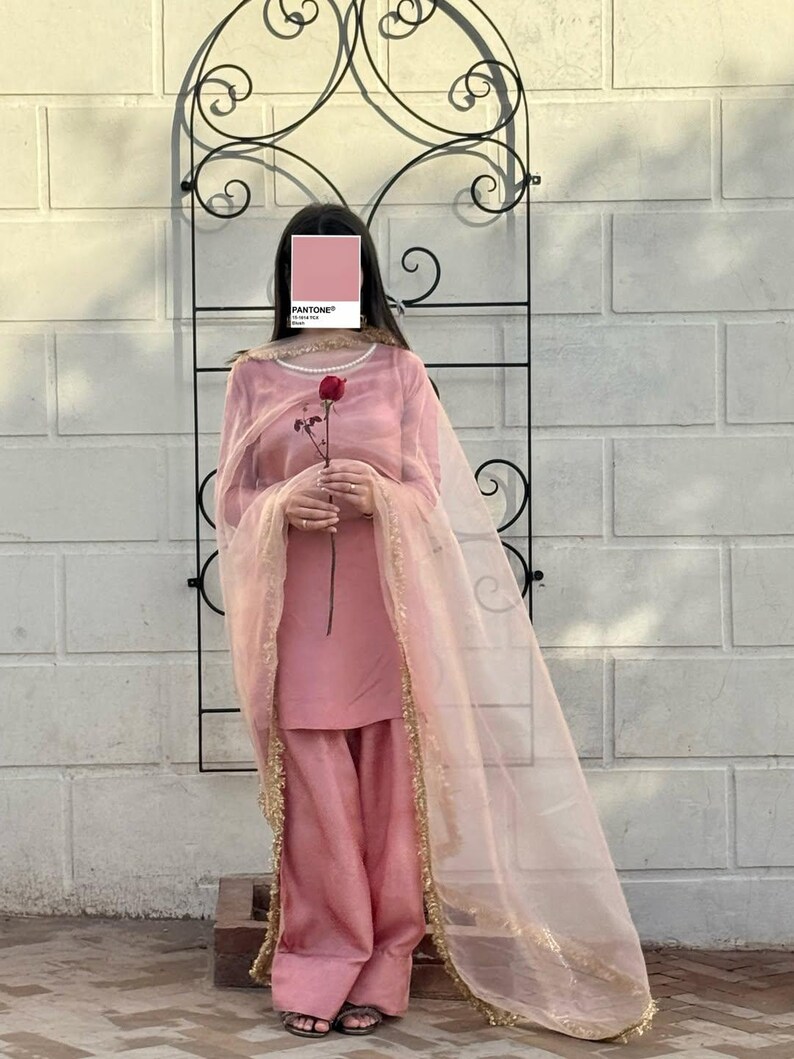 May include: A light pink salwar kameez outfit. The ensemble includes a long-sleeved tunic, wide-legged pants, and a sheer dupatta with gold trim. A red rose is held in the hands. The background features a decorative black metal arch and a white brick wall.
