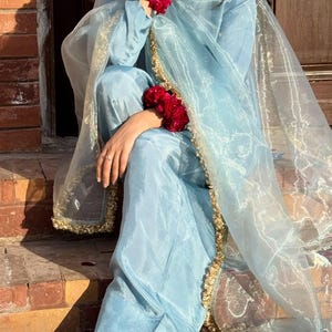 May include: A light blue silk outfit, including a long-sleeved top and pants, is accented with gold trim and a sheer scarf. The outfit is adorned with red floral wristlets. The shoes are gold with a floral design.
