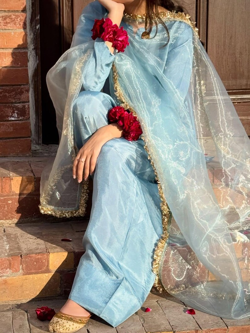 May include: A light blue silk outfit, including a long-sleeved top and pants, is accented with gold trim and a sheer scarf. The outfit is adorned with red floral wristlets. The shoes are gold with a floral design.
