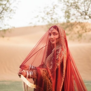 Puede incluir: Una mujer con un vestido de novia rojo tradicional con bordados dorados y un velo transparente. El velo está drapeado sobre la cabeza y los hombros, y el vestido presenta detalles intrincados. El fondo muestra un paisaje desértico.