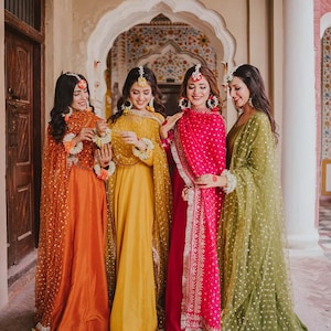 Op de afbeelding: Vier vrouwen in kleurrijke traditionele kleding poseren buiten. De jurken zijn in tinten van oranje, geel, roze en groen. Elke vrouw draagt bijpassende hoofddeksels en sieraden. De setting is een stenen boog met sierlijke details.