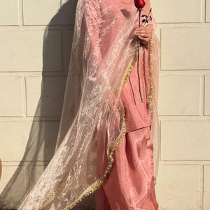 May include: A light pink salwar kameez ensemble. The outfit includes a tunic, loose-fitting pants, and a sheer dupatta. The dupatta is trimmed with gold detailing. A red rose is held in the hand. The outfit is worn in front of a white brick wall.