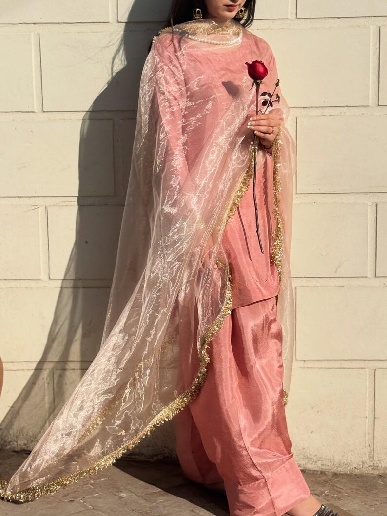 May include: A light pink salwar kameez ensemble. The outfit includes a tunic, loose-fitting pants, and a sheer dupatta. The dupatta is trimmed with gold detailing. A red rose is held in the hand. The outfit is worn in front of a white brick wall.