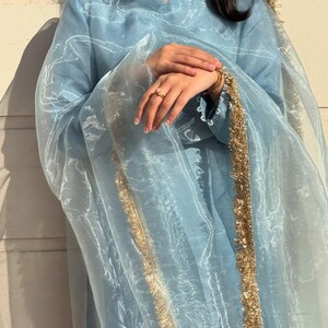 May include: A light blue ensemble featuring a long-sleeved tunic and a sheer scarf, both adorned with gold trim. The outfit includes gold jewelry and a ring. The fabric has a subtle sheen, creating an elegant look.