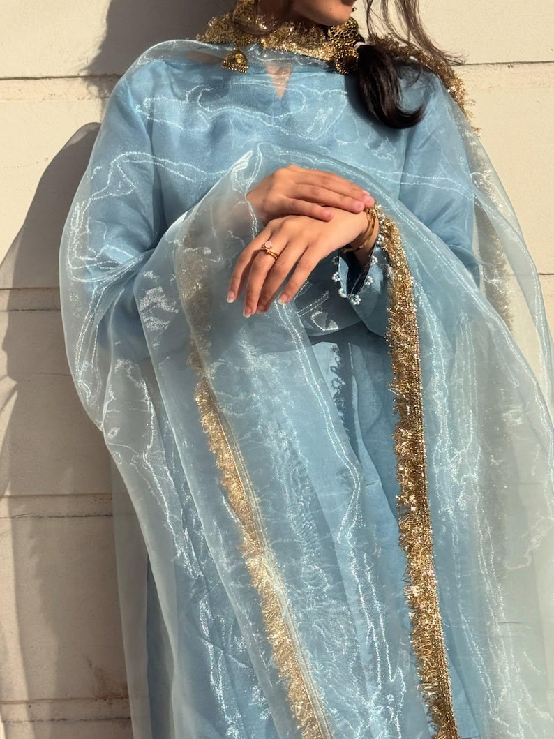 May include: A light blue ensemble featuring a long-sleeved tunic and a sheer scarf, both adorned with gold trim. The outfit includes gold jewelry and a ring. The fabric has a subtle sheen, creating an elegant look.