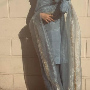 May include: A light blue ensemble featuring a long-sleeved top and matching pants, complemented by a sheer, flowing scarf. The scarf and the edges of the outfit are adorned with gold trim, creating a delicate and elegant look.