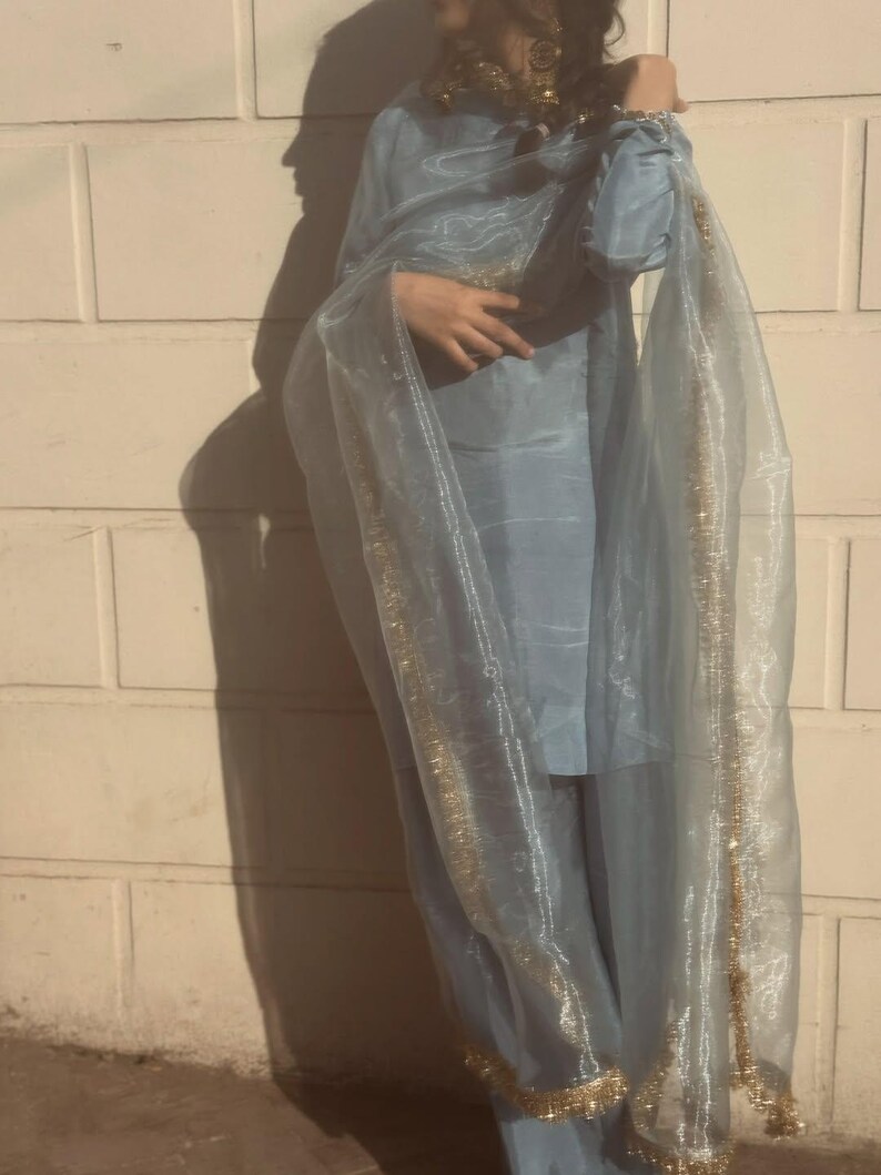 May include: A light blue ensemble featuring a long-sleeved top and matching pants, complemented by a sheer, flowing scarf. The scarf and the edges of the outfit are adorned with gold trim, creating a delicate and elegant look.